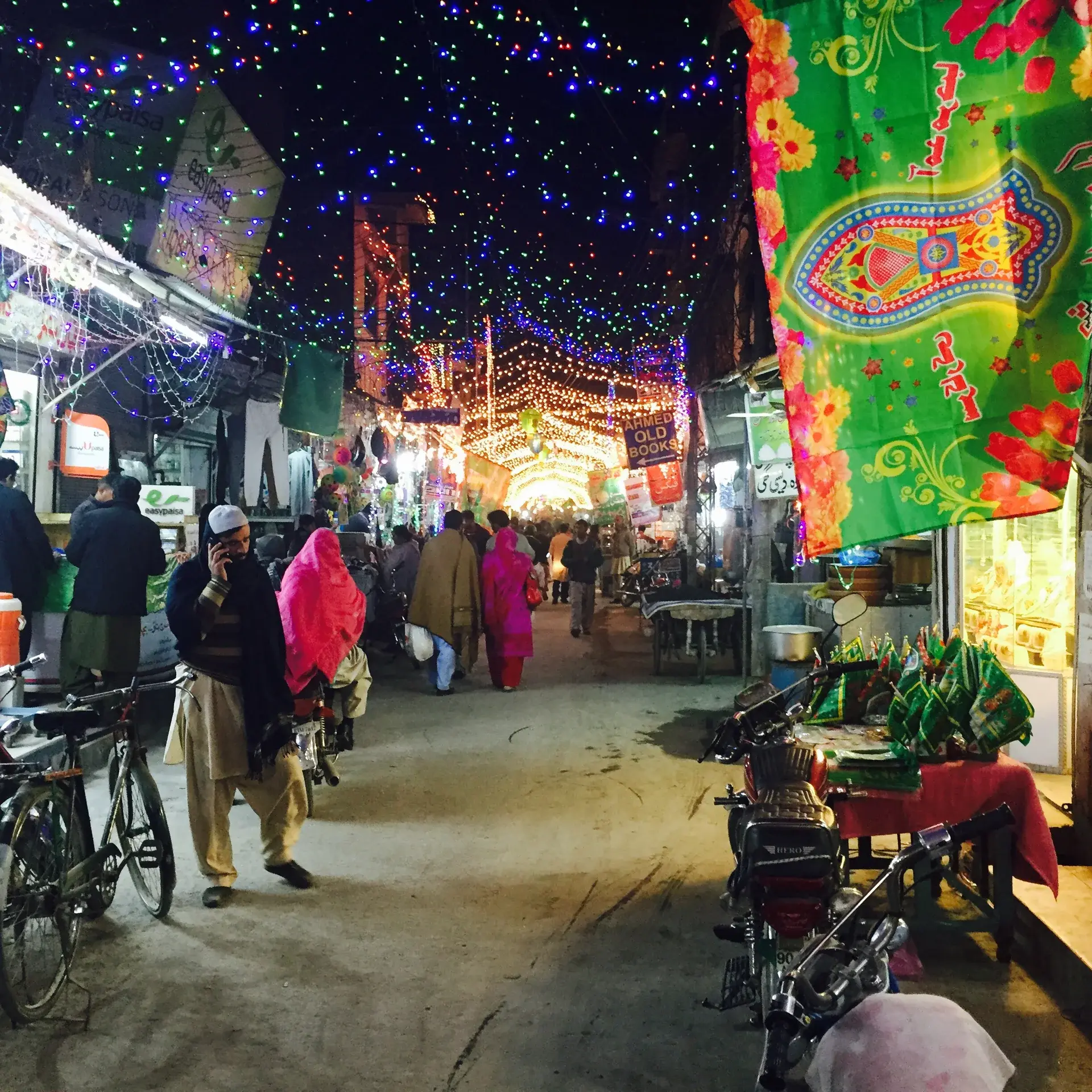 People walking between shops and market stalls beneath colourful evening lights in Lalkurti, Rawalpindi